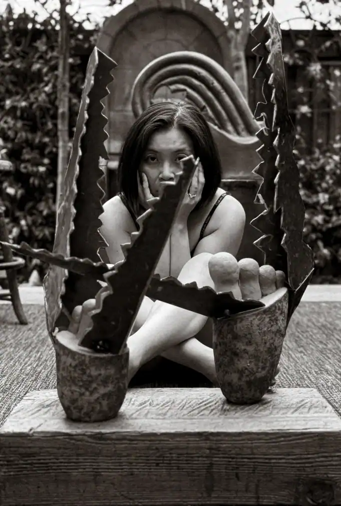 Photograph of Loni Stark seated behind her sculpture CrocodIle (2025), which features jagged steel jaws and rounded ceramic toes mounted on a wooden base.
