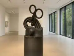 Bronze sculpture featuring a large pair of scissors embedded in abstract loops and curves, displayed in a minimalist gallery with floor-to-ceiling windows.
