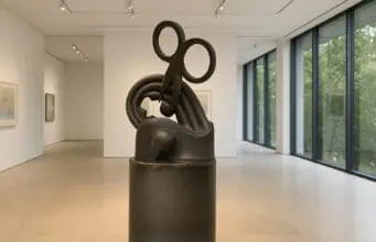 Bronze sculpture featuring a large pair of scissors embedded in abstract loops and curves, displayed in a minimalist gallery with floor-to-ceiling windows.