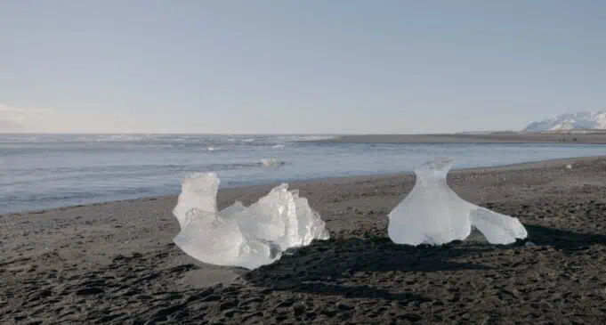 Melting glacier ice chunks on dark sand beach in Greenland with mountains in background