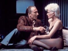 Scene from Brian De Palma’s Body Double (1984) with a woman in black lingerie talking to a man in a leather jacket on a bed in a Hollywood apartment high above Los Angeles, California.