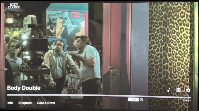 Screenshot of Body Double (1984) showing a film camera crew reflected in a doorway during the Holly Body sequence, with leopard print wall and neon colors on a Hollywood set in Los Angeles, California.