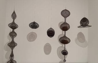 Six Ruth Asawa looped-wire sculptures suspended against white wall with circular shadows